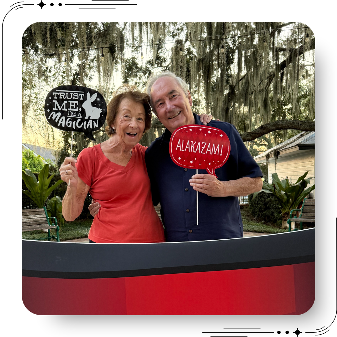 Smiling couple posing with “Trust Me I’m a Magician” and “Alakazam” signs at a Mount Dora Florida magic event, outdoor festival setting with Spanish moss trees in background.