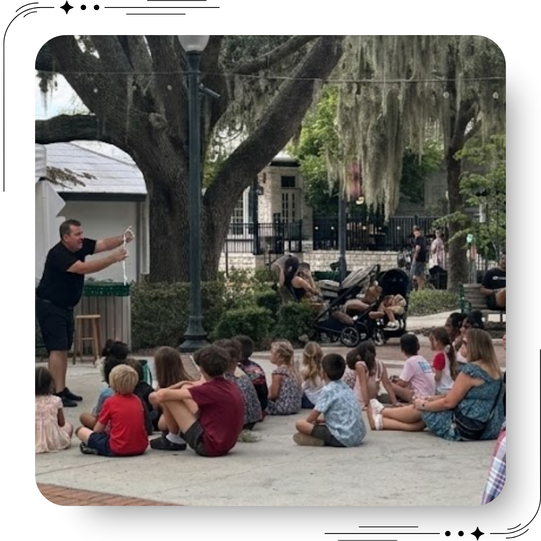 Children and families watching a live outdoor magic show under oak trees in downtown Mount Dora Florida, community festival atmosphere with parents and kids gathered in the park.